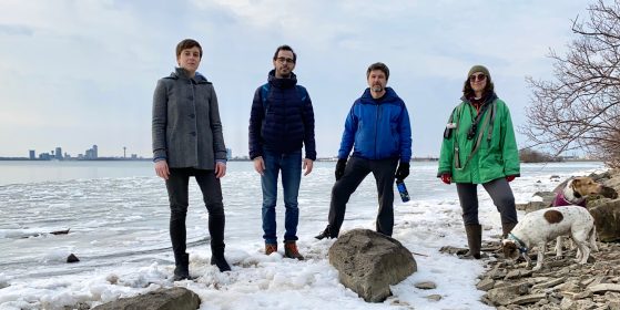 The four members of The Evolution of the Arm standing together in the ice on a beach with Toronto on the other side of the water.