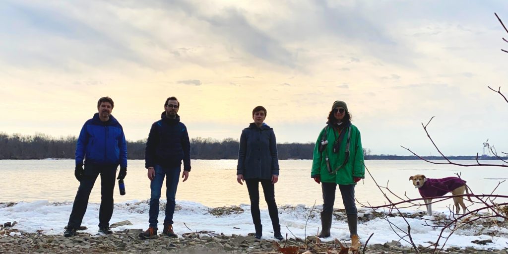 The four members of The Evolution of the Arm standing together in the ice on a beach with Toronto on the other side of the water.